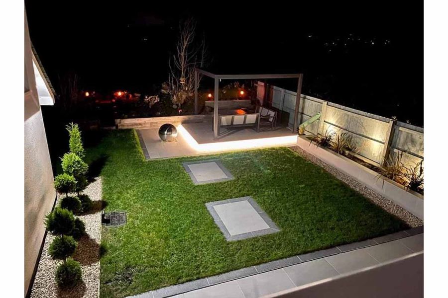 Garden illuminated at night, with large stepping stones in lawn and patio paving edged with Charcoal porcelain planks.