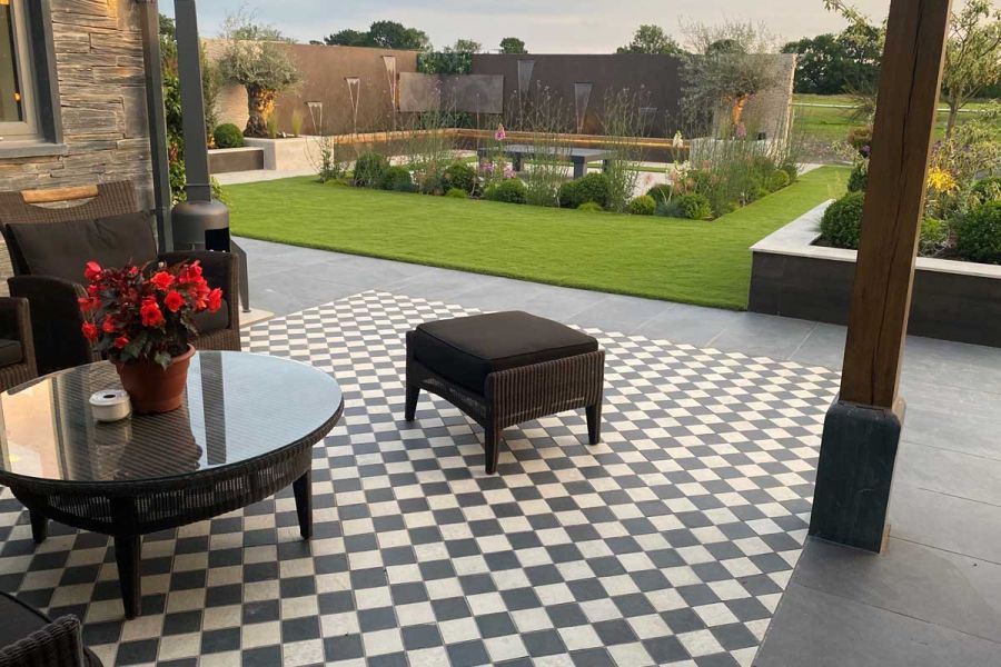 Lawn and wall water feature beyond patio with chequerboard layout of small porcelain patio tiles in light grey and charcoal.