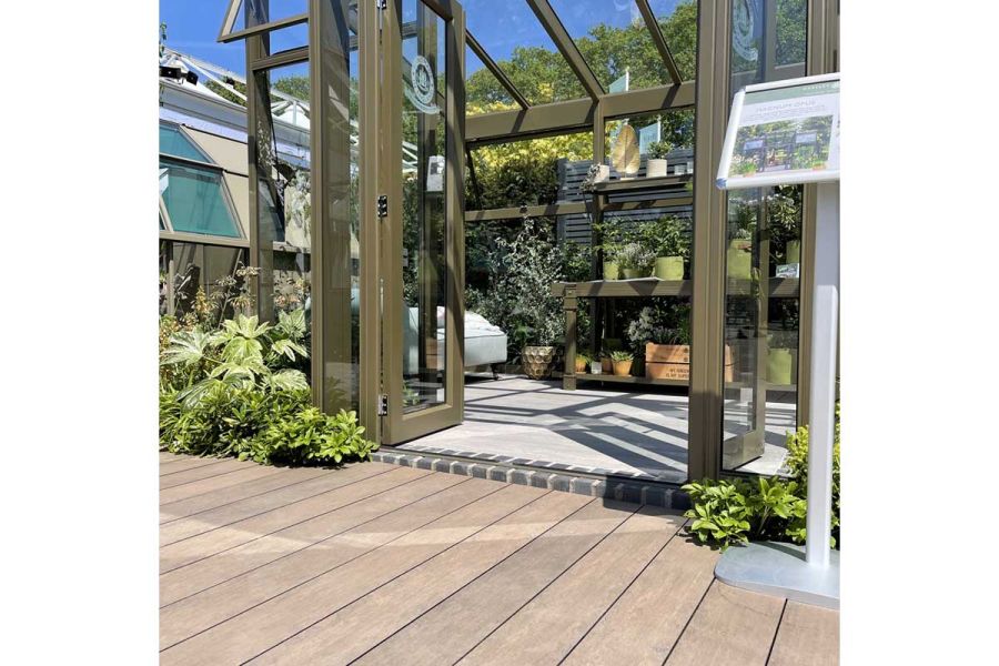 Metal greenhouse with staging, plants and chair in front of Chestnut Brushed DesignBoard decking. Trade stand designed by Llevelo.