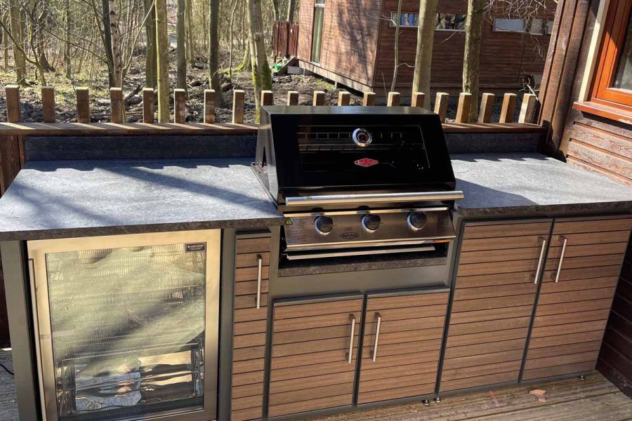 Outdoor kitchen set against fence at edge of decking, with cooler and units faced with Chestnut composite battens. Woodland beyond.