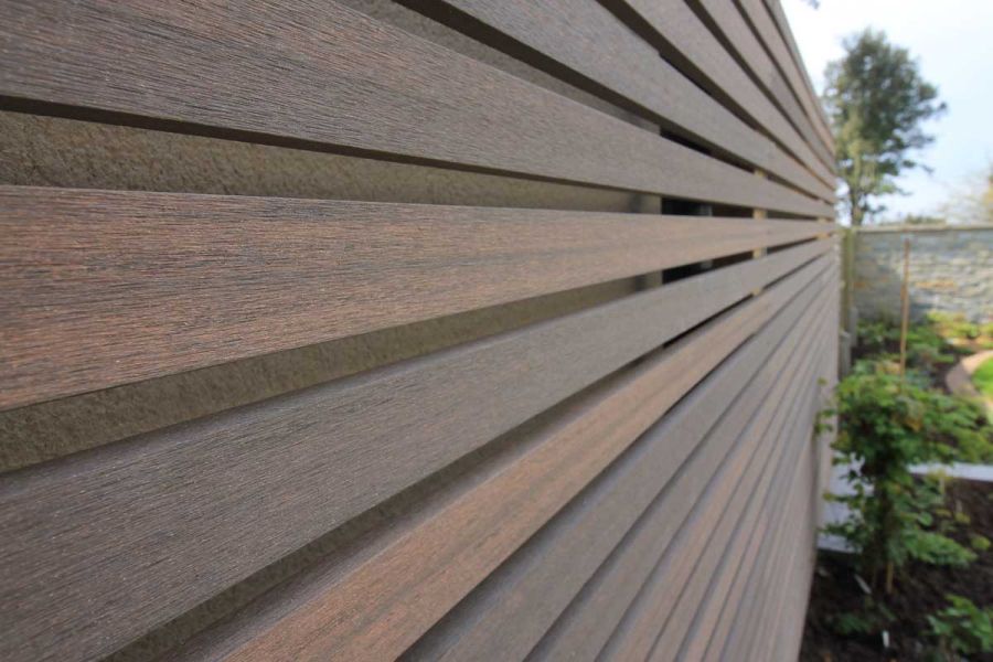 Angled view along open-slatted screen of Chestnut composite battens. Planted bed below. Stone clad wall in distance.