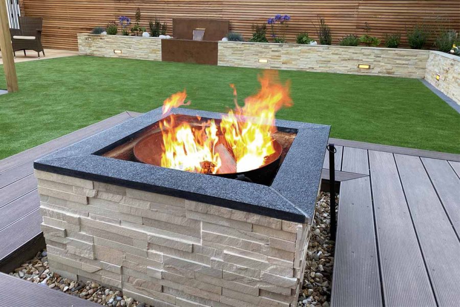 Stone-clad square firepit with granite coping set into Chestnut Grooved composite decking in front of lawn edged with raised beds.