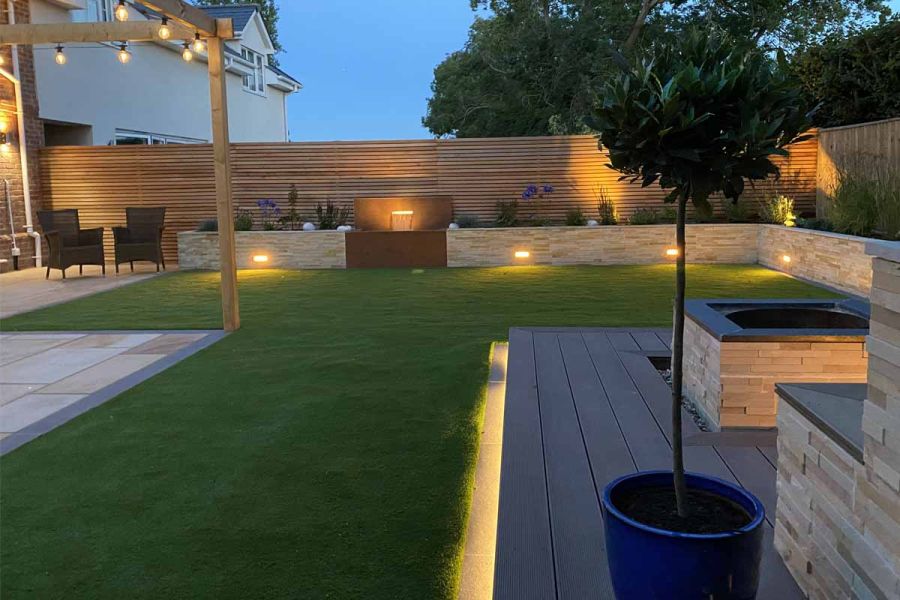 Deck of Chestnut Grooved DesignBoard with large lawn edged with stone-clad raised beds and slatted fence, illuminated at dusk.