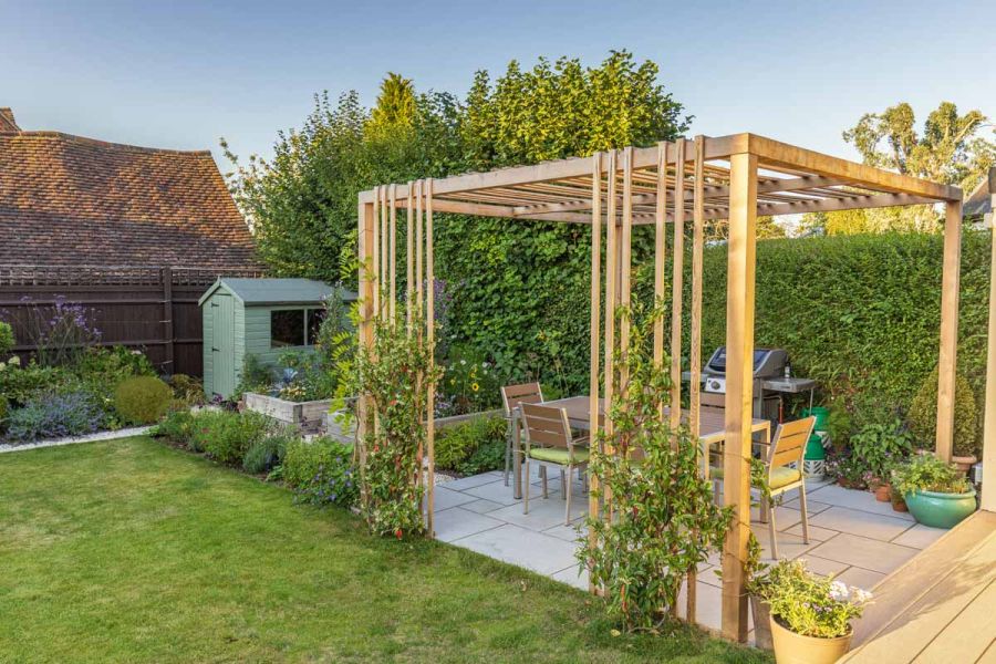 Modern wooden pergola stands on patio paved with Dove Grey smooth sandstone project pack slabs in hedged garden with lawn and shed.