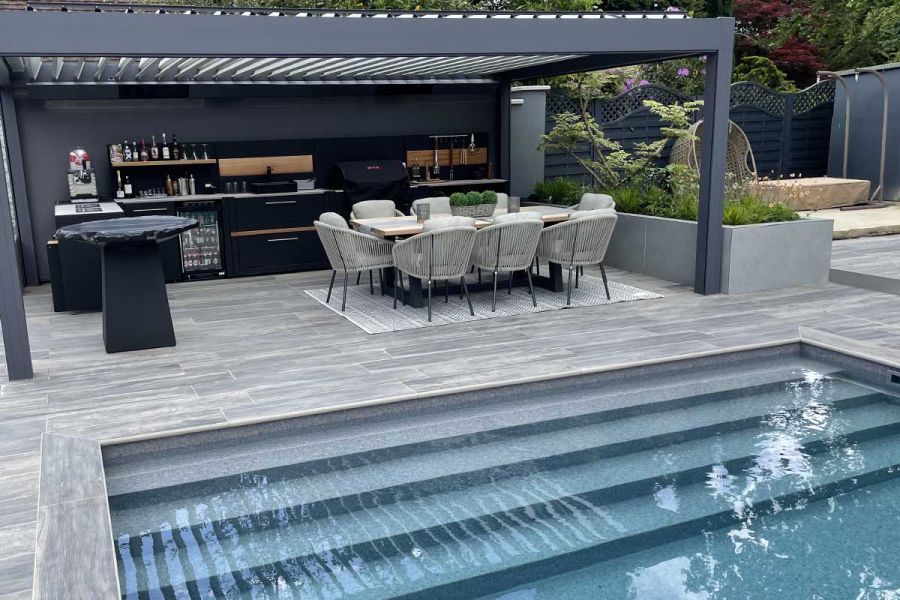 Patio with pergola, outdoor kitchen and dining set. Cinder bullnose porcelain steps make edge between paving and swimming pool.