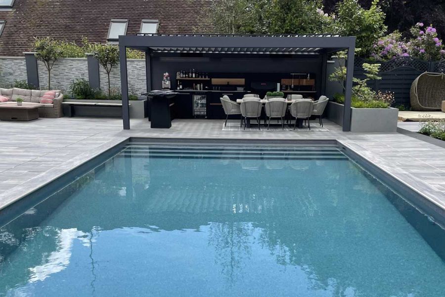 Swimming pool with Cinder porcelain bullnose steps used as coping, surrounded by matching patio paving in large garden with pergola.