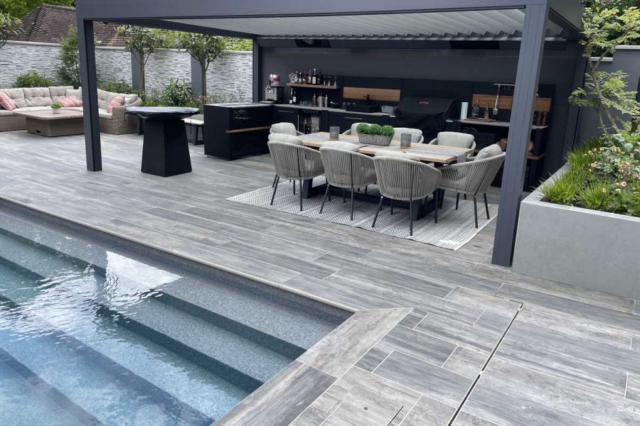 Cinder porcelain bullnose step treads edge swimming pool set into patio of matching paving, with outdoor kitchen under pergola.