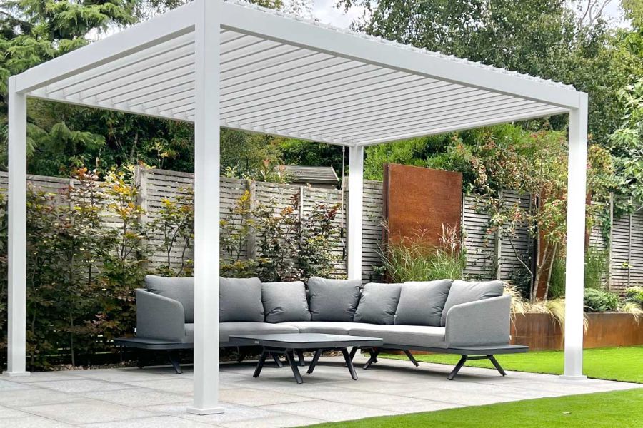 Large Proteus White aluminium weatherproof pergola over sofa on rectangular paved area in garden with fence and planting behind.
