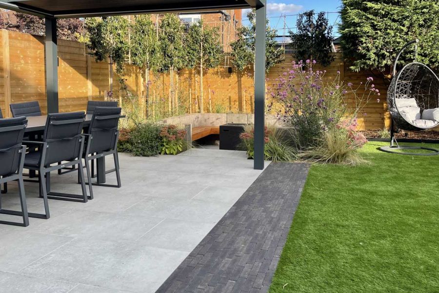 Pergola-covered patio edged with Amersham brick pavers leads to seating area within built benches in fenced garden with lawn.