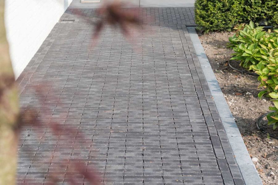 Wide path of Carbona Dutch clay pavers laid in 2 directions runs between white wall and with grey kerb down to bed with shrubs.