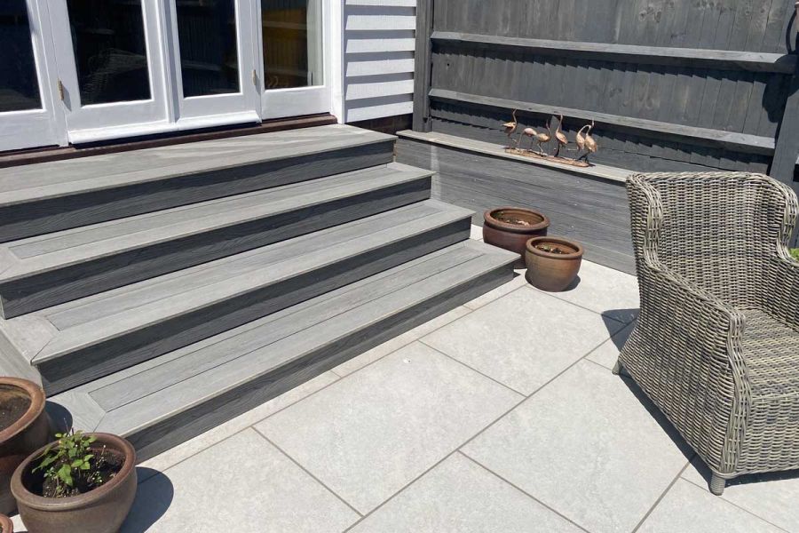 Grey decking steps leading down from white French doors. Astor Grey Porcelain Paving with planters and rattan armchair on the patio.
