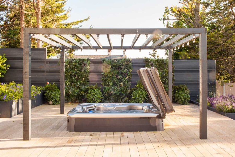 Open hot tub under pergola with lights over Cinnamon composite decking. Slatted fence behind. Design by Simon Orchard.