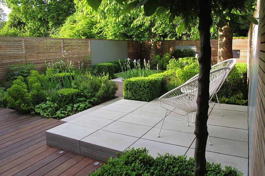 Raised square of Contemporary Grey sawn sandstone paving laid stack bond, in fenced garden with low hedges edging and decking.