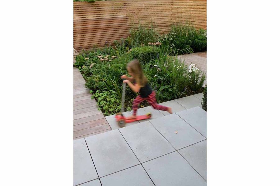 Child on scooter races along path past raised Contemporary Grey patio and rectangular flower bed. Design by Lucy Wilcox.