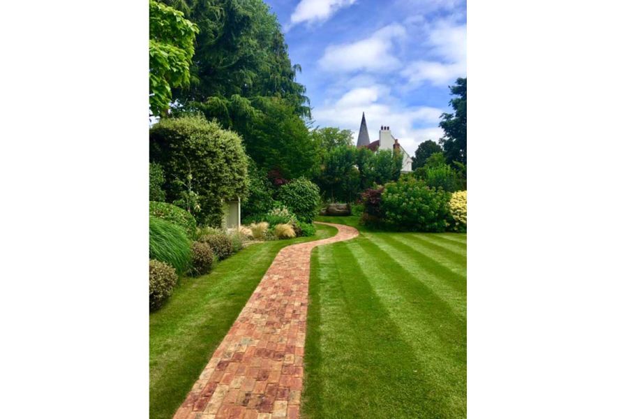Straight path of Cotswold Blend Clay paver bricks cuts through striped lawn by lushly planted border and curves between bushes.