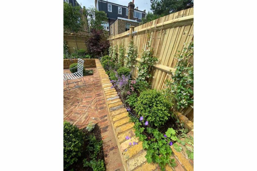 View along garden fence and brick-built raised bed bordering Cotswold clay paver patio with a metal chair and beds set into paving.