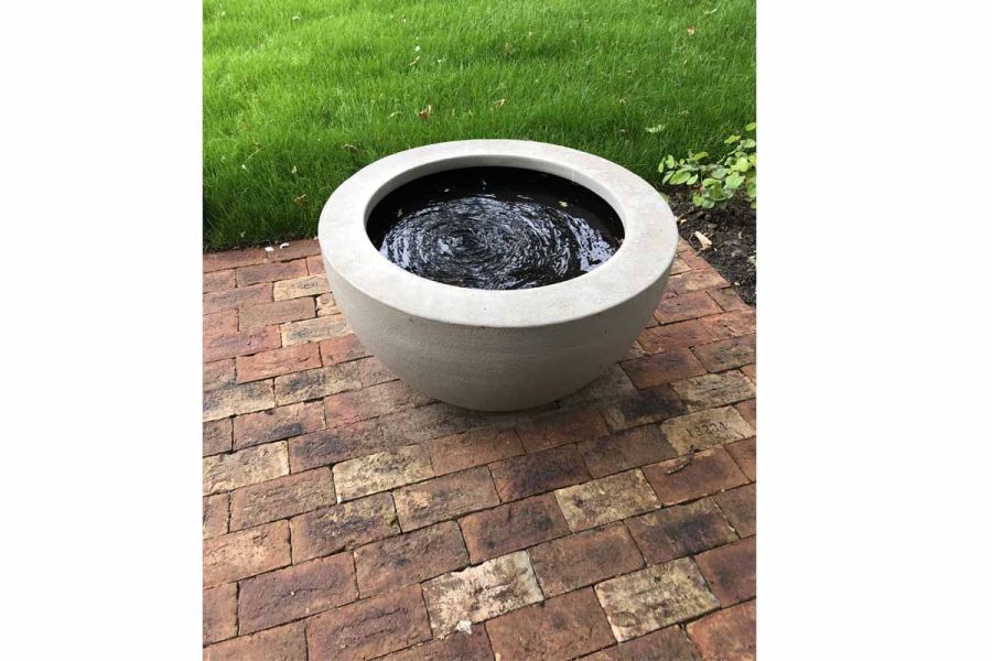 Round concrete bowl water feature sits on paved area of Cotswold clay bricks laid flat next to lawn and flower bed.