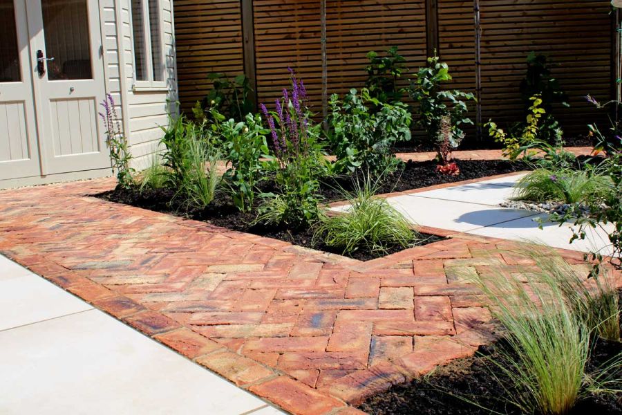 Cotswold clay pavers laid herringbone with inset beds between cream paving in front of garden house. Built by Essex Garden Designs.