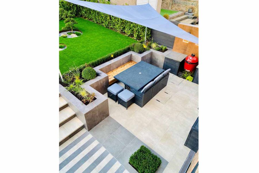 View down on patio of cream porcelain plank paving and 900x600mm slabs with steel-grey paving. Steps rise from dining area to lawn.