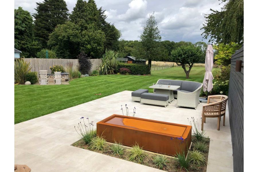 Garden furniture and corten steel water feature sitting amidst a cream porcelain patio at the top of large garden backing onto meadows.