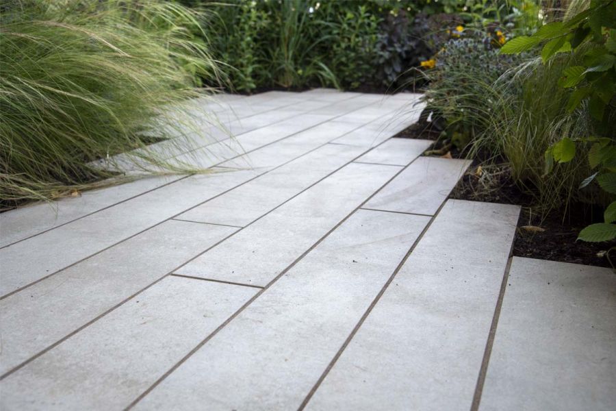 Cream porcelain planks laid between planted borders, seen from low angle. Ends of plank paving create stepped edge to beds.