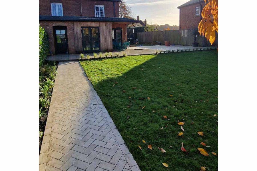 Path of Cream porcelain setts leads down side of lawn to back of house with wood-clad extension and patio wrapped around side.