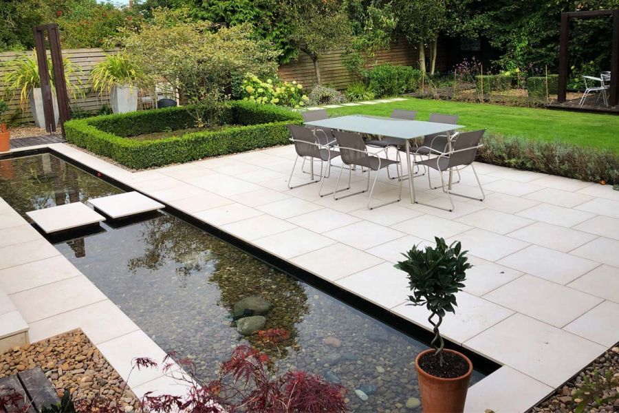 Oblong pond, with 2 Florence White outdoor tile stepping stones. Matching patio next to hedged lawn. Design by Creative Roots.