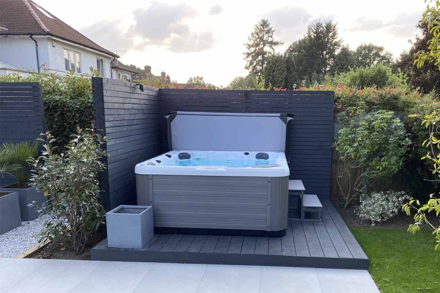 Grey-sided hot tub stands on raised deck of Dark Ash Brushed outdoor composite decking, backed on 2 sides by slatted fencing.