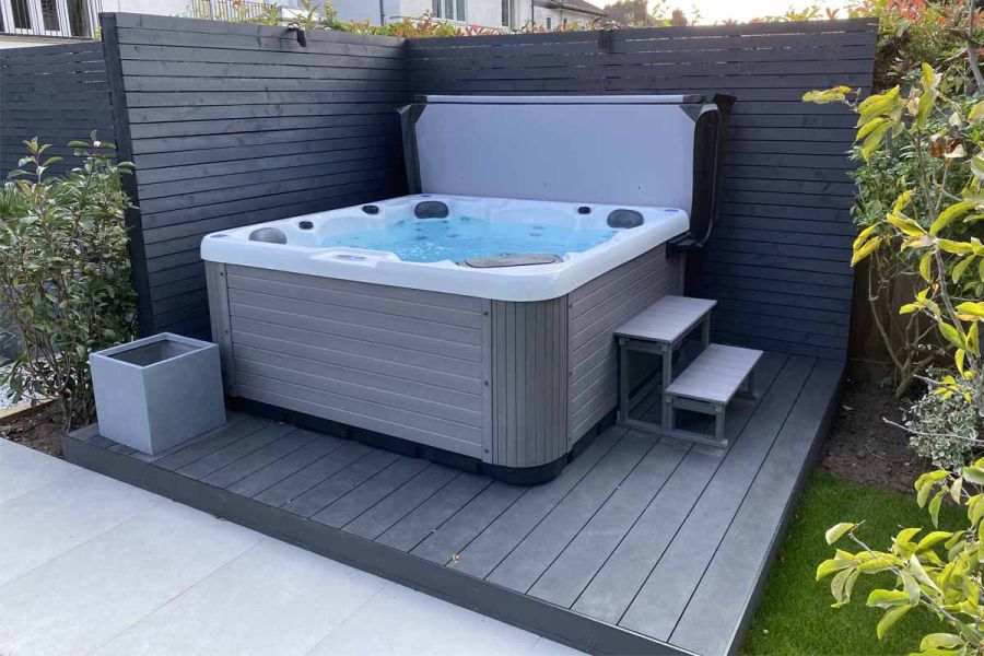Hot tub on raised platform of Dark Ash Brushed plastic decking with matching fascia board. Slatted grey fence on 2 sides.