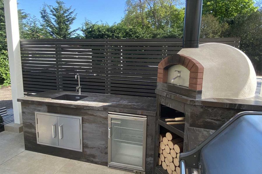 Outdoor kitchen with fridge and wood-burning pizza oven, and sink unit backed by screen of Dark Ash composite battens.