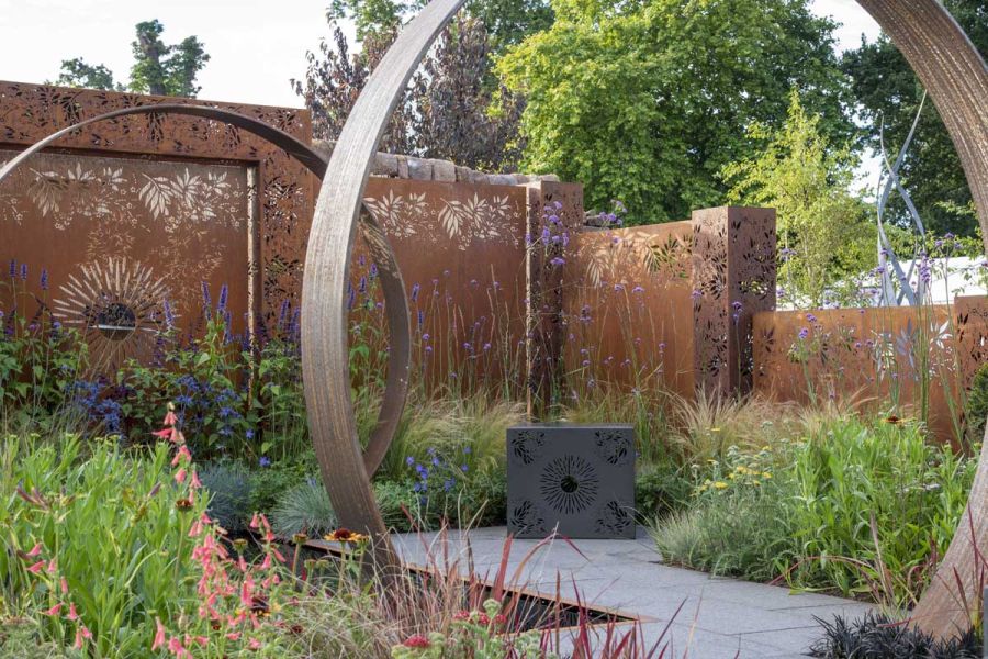 Metal pierced screens fence RHS show garden by Charlie Bloom with Dark Grey Granite paving, hoop arches and lush planting.