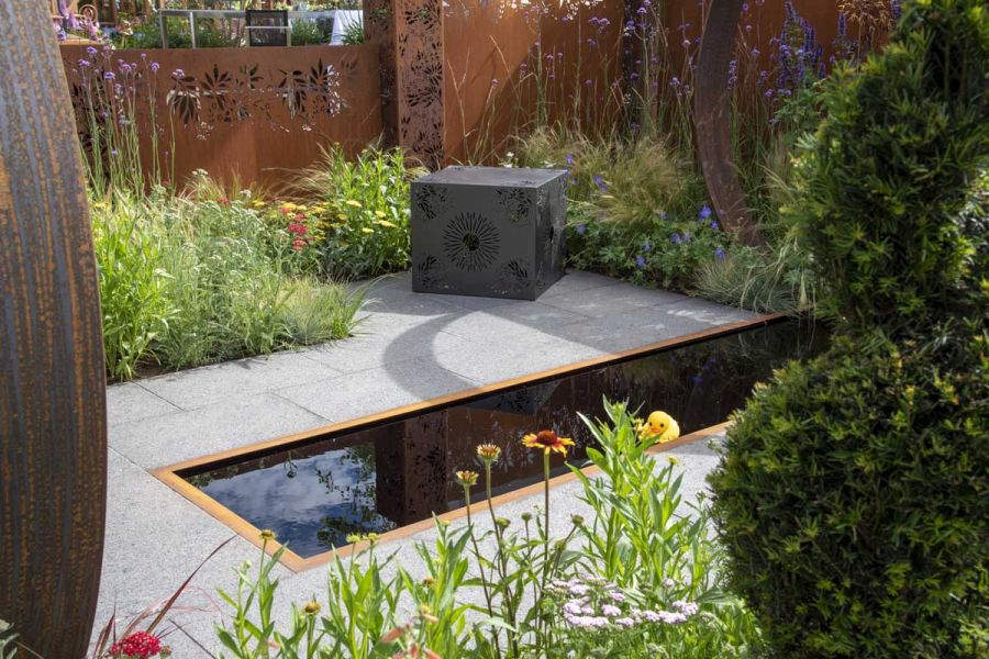 Metal cube seat on Dark Grey Granite paving, in RHS show garden by Simon Webster, with oblong pond and metal fencing.