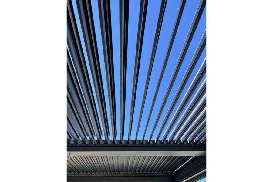 View up at roof of Dark Grey Metal Pergola. Louvres are open in one half, showing blue sky beyond, and the rest are closed.