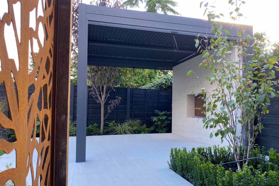 Dark Grey Metal Pergola placed over cream paving with matching wall at back. Oxidised metal screen and formal bed in foreground.
