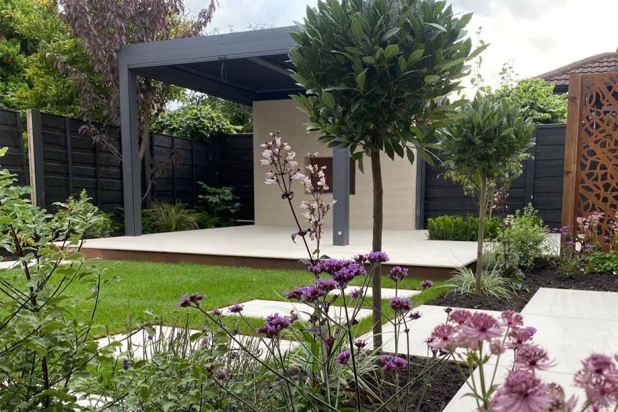 Dark Grey Metal Pergola over raised cream-paved patio, with lawn and flower beds. Design by Signature Landscapes Design and Build.