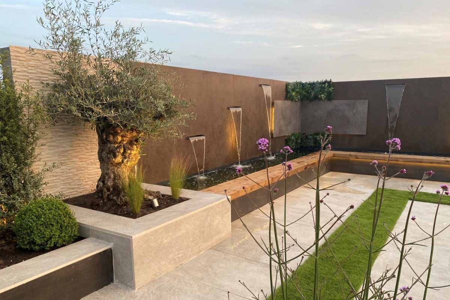 Boundary wall faced with Dark Mocha DesignClad, with 3 water spouts emptying into pond behind bench-top wall, next to olive tree.