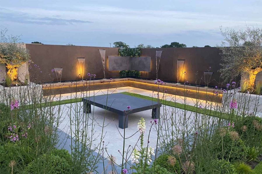 Low square table sits on paved area edged by beds and an L-shaped pond backed by wall of Dark Mocha exterior cladding.