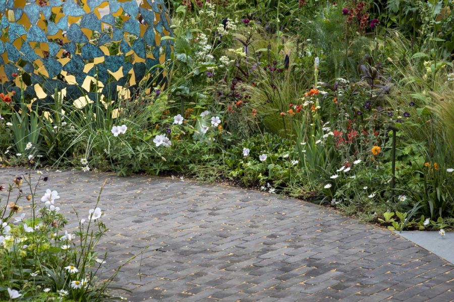 Curved path of Lucca brick paving between lushly planted beds with round pierced sculpture on David Harber RHS Chelsea trade stand.