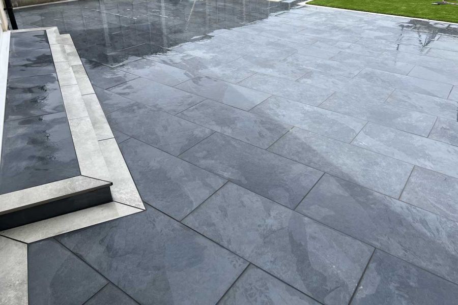 Large wet patio of Slab Coke 900x600 porcelain slabs with 2 steps edged in light grey planks. Built by Delta Landscape Solutions.
