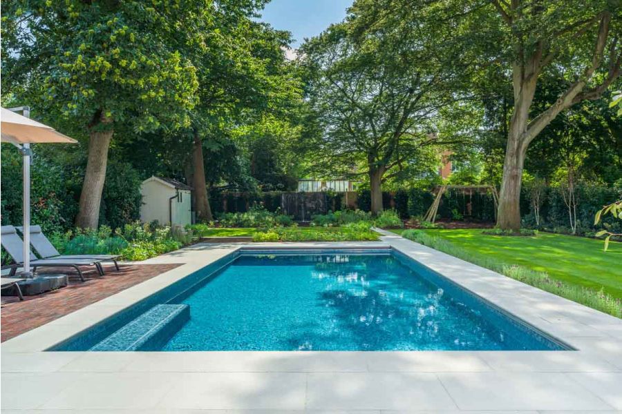 Swimming pool in garden with mature trees and lawns. Pool surround and paths of Dove Grey Sawn sandstone paving, with clay pavers.