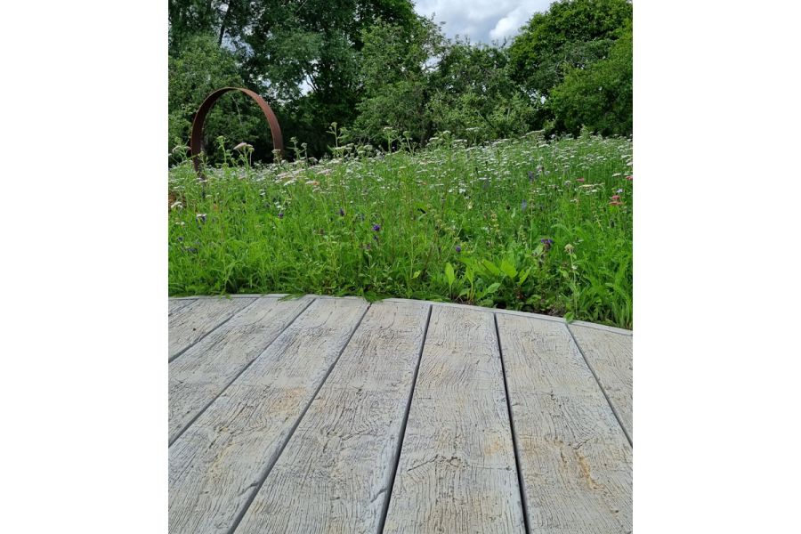 Low view of grey Driftwood Millboard decking with curved edge. Natural planting and Corten steel circular sculpture in background. 