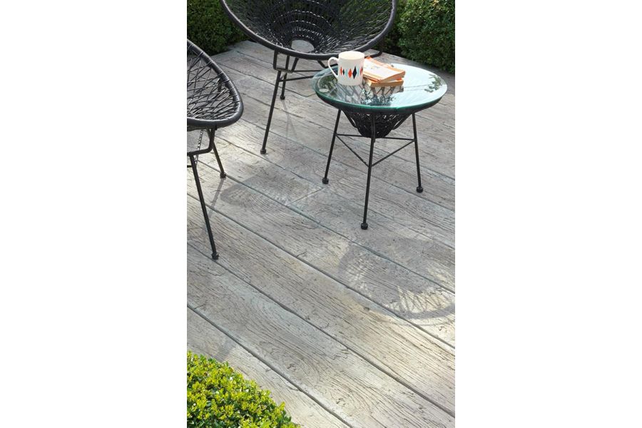 2 black bucket chairs and table cast shadows on Driftwood Millboard decking edged with low trimmed hedge.