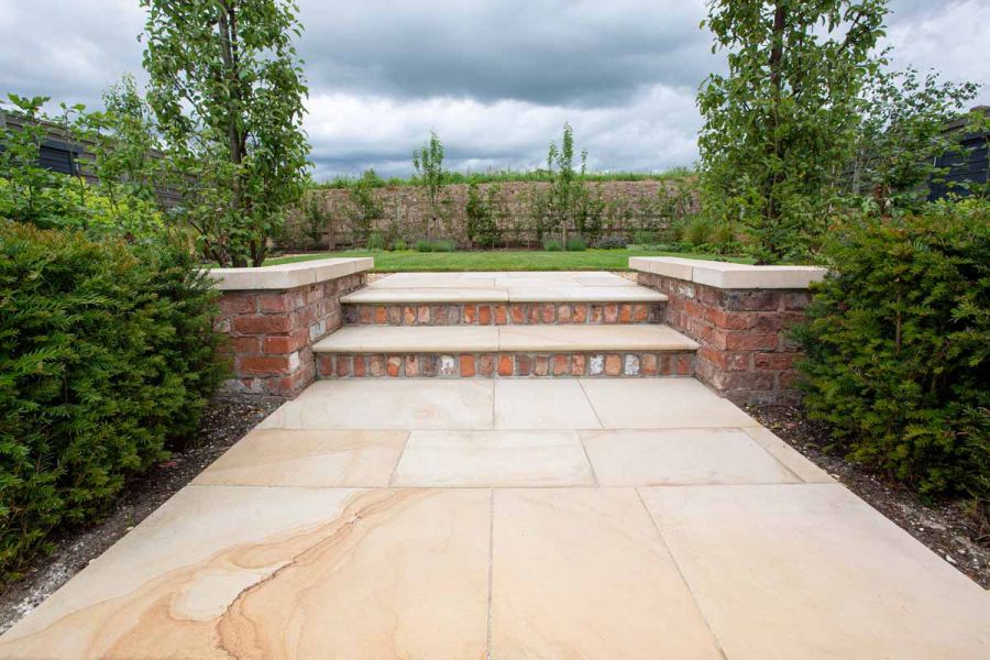 2 Dune Sawn Sandstone steps with brick risers and retaining walls topped with thick Dune coping slabs rise from path to lawn.
