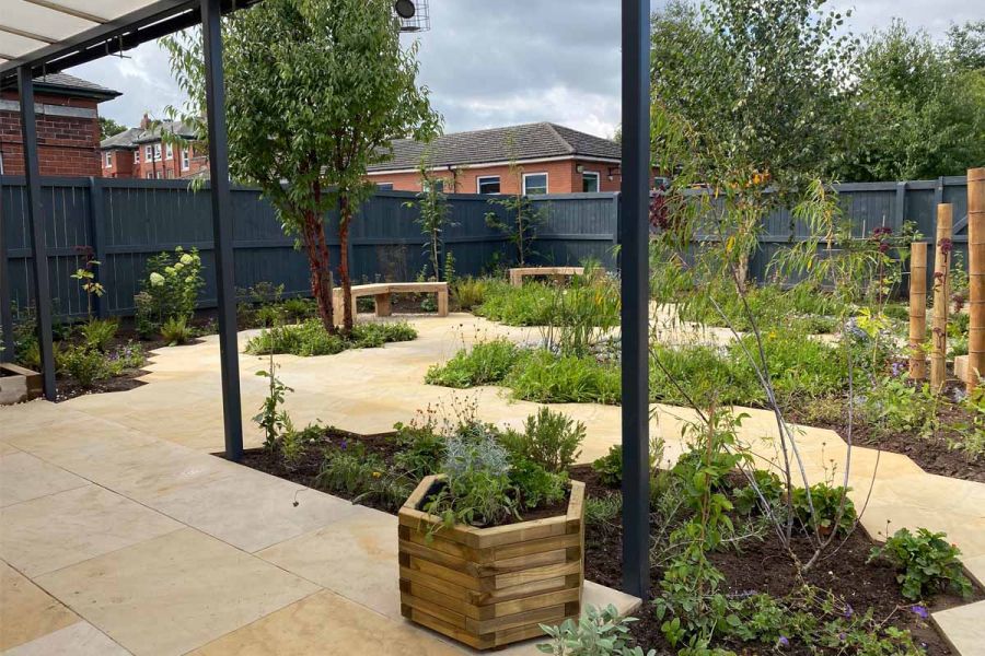 Grey-fenced garden, paved in Dune Smooth Sandstone, with planted beds inset into paving, wooden benches and octagonal planters.