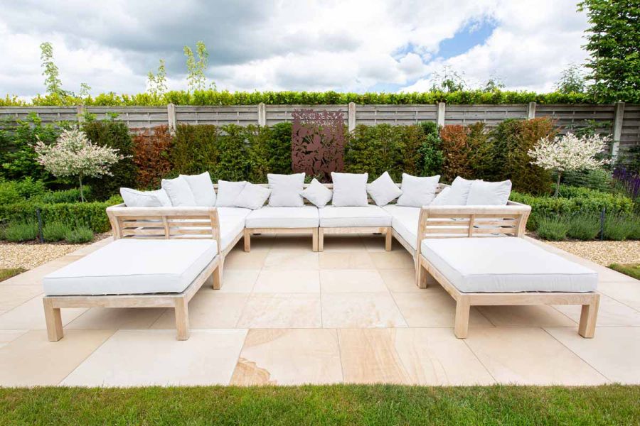 Large modular, white-cushioned luxury wooden furniture on Dune Smooth Sandstone paving slabs. Fence and hedged flower bed behind.