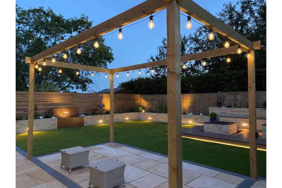 Wooden pergola hung with lights stands over rectangle of Dune sawn sandstone paving set into corner of lawn edged with raised beds.