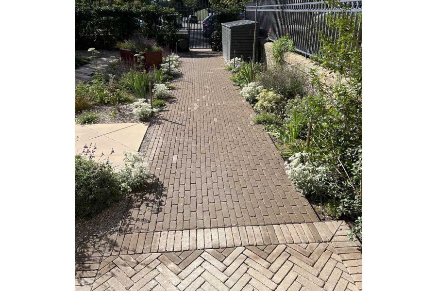 Wide path of Aldridge clay pavers leads between wall with railings and plant borders to tall iron gate set into hedge.