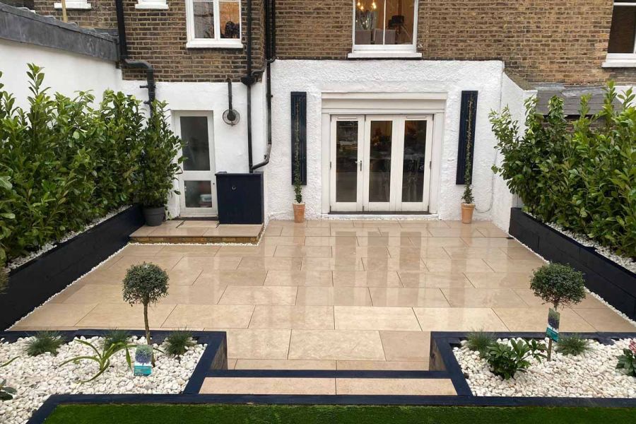 Straight-sided, long rectangular back garden entirely paved in Egyptian Beige Limestone paving, edged with shrubs and trees.
