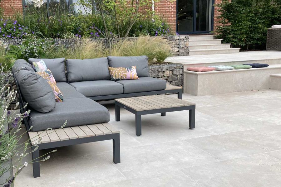 Modular sofa and table sit in corner of patio paved with Egyptian Beige limestone bordered by stone-filled gabions and grasses.