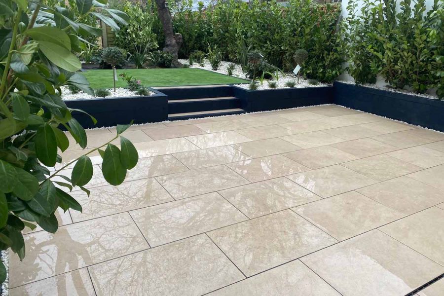 Large patio of wet Egyptian Beige Limestone with steps between retaining wall and gravelled beds up to lawn edged with shrubs.